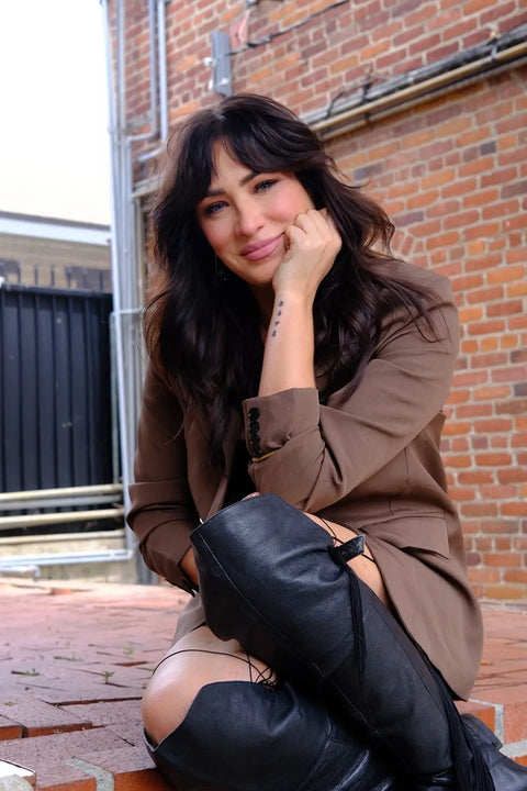 A woman with dark hair sits on brick steps against a red brick wall, wearing a brown blazer, a light beige top, and black leather knee-high boots.